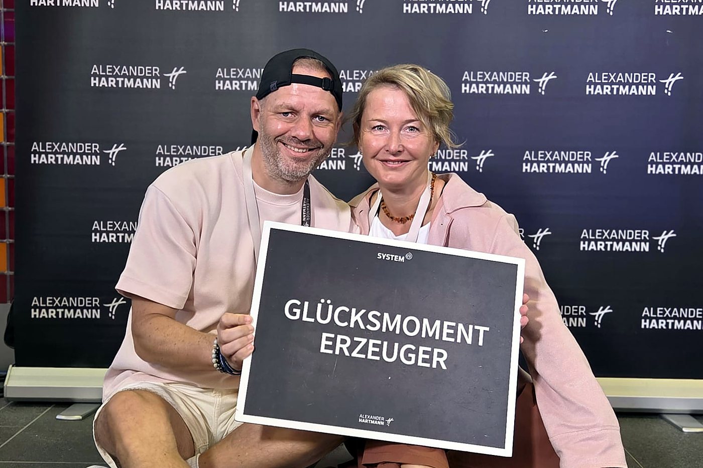 Ralf Hermann und Nicole Butschbach sitzen lachend vor einer Fotowand und halten ein Schild mit der Aufschrift „Glücksmoment-Erzeuger“
